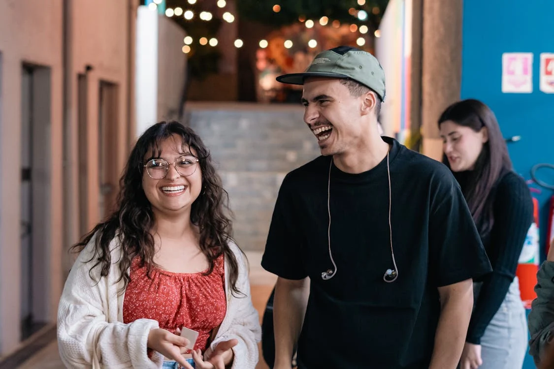 Two young people smiling standing side by side. On the left is a woman with glasses and curly hair. on the right a man with a hat and earphones on his neck